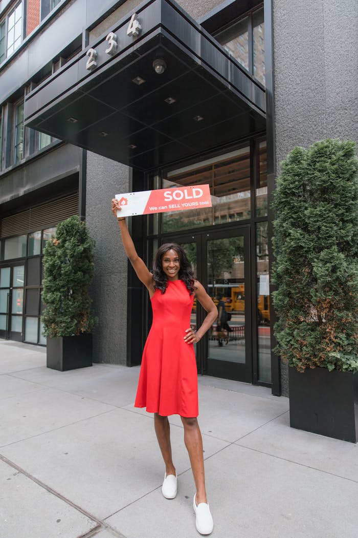 Crafting Captivating Headlines: Your awesome post title goes here Smiling woman in red dress holding a sold sign outside a building, showcasing real estate success.