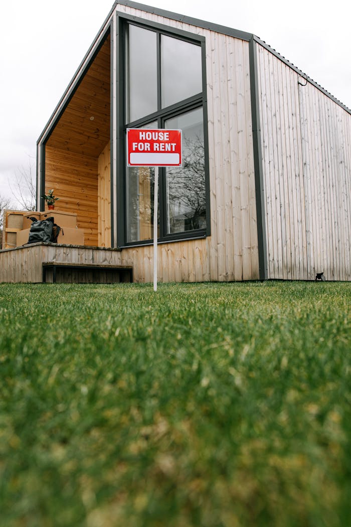 A modern wooden house with a 'for rent' sign on a lush green lawn.